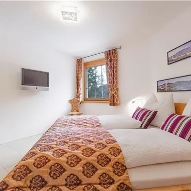 Ein gemütliches Schlafzimmer mit zwei Betten und bunten Kissen. Das Zimmer hat ein Fenster mit Blick auf die Bäume und ein modernes TV-Gerät an der Wand.