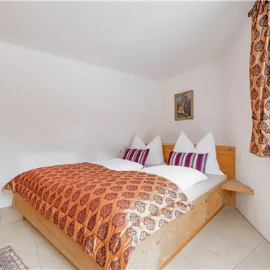 Ein gemütliches Schlafzimmer mit einem Doppelbett und farbenfrohen Kissen. Die Wände sind schlicht und es gibt einen großen Fenster mit Vorhängen.