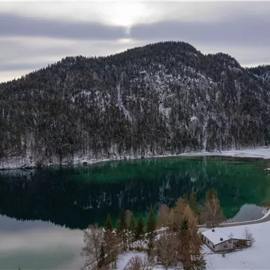 Ein malerischer See umgeben von schneebedeckten Bergen und Wäldern. Die ruhige Wasseroberfläche spiegelt die natürliche Schönheit der Umgebung wider.