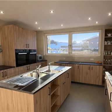 A modern kitchen with wooden cabinets and simple gray countertops. Large windows provide plenty of daylight.