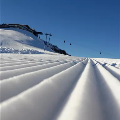 Eine beeindruckende verschneite Landschaft mit perfekt präparierten Schneepisten. Im Hintergrund sind kabelgebundene Lifte und ein klarer blauer Himmel zu sehen.