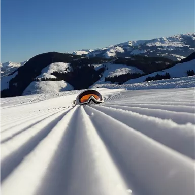 Eine schneebedeckte Landschaft mit Bergen im Hintergrund. Eine Skibrille liegt im Schnee und die Sonne scheint klar am blauen Himmel.