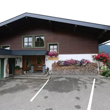 Ein elegantes Holzhaus mit einem großen Dach und bunten Blumen vor der Fassade. Im Vordergrund befinden sich Parkplätze und eine grüne Landschaft im Hintergrund.