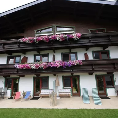 Ein modernes Haus mit mehreren Balkonen, die mit bunten Blumen dekoriert sind. Vor dem Gebäude befinden sich Sitzgelegenheiten und Liegen im Garten.