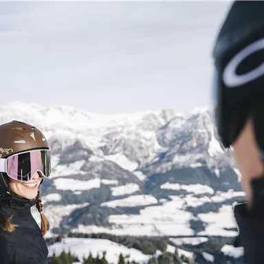 Two skiers are smiling at each other on a snow-covered mountain. In the background, majestic mountains and a clear sky can be seen.
