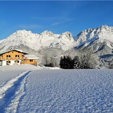 A cozy house is situated in a snow-covered landscape. In the background, majestic mountains rise under a blue sky.