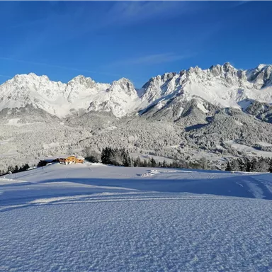 A snow-covered landscape with majestic mountains in the background. The sky is clear and blue, creating a serene winter atmosphere.