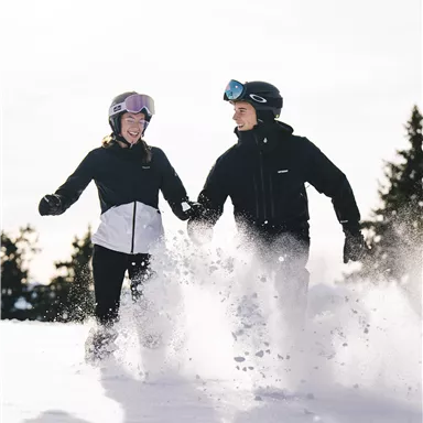 A couple in the snow is having fun and playing together. Both are wearing ski gear and laughing as they walk through the fresh snow.