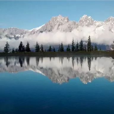 A clear lake reflects majestic mountains and a blue sky. Surrounded by green trees and light mist.