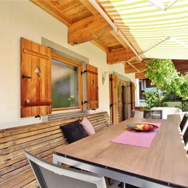 An inviting outdoor area with a large dining table and wooden benches. The sunshade and the green garden create a relaxed atmosphere.
