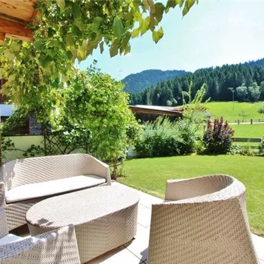 An inviting garden with cozy rattan seating. In the background, green meadows and wooded hills can be seen.