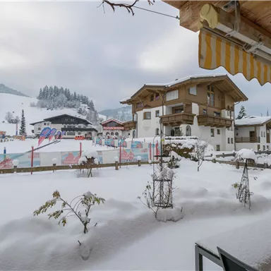 A winter landscape with a lot of snow and a view of traditional alpine houses. In the background, snow-covered mountains and a ski resort can be seen.