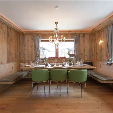 A cozy dining area with wooden paneling and a large table decoration. The green chairs and the chandelier give the room an inviting atmosphere.