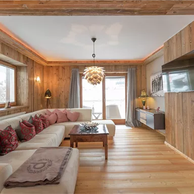 A cozy living room with wooden walls and a large, comfortable sofa. The room is well-lit and features a modern TV unit, creating a pleasant atmosphere.