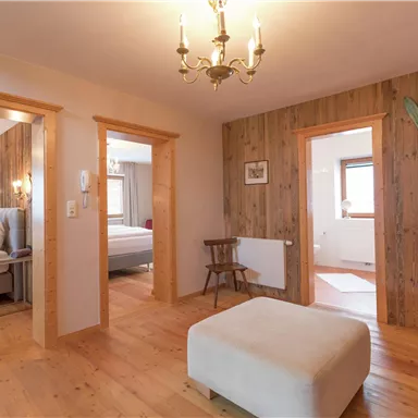 A cozy room with wooden walls and a modern chandelier. In the background, two bedrooms and a seating area can be seen.