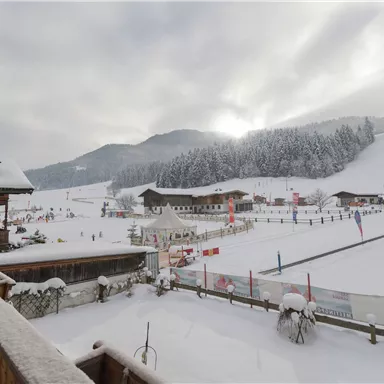 A winter landscape with plenty of snow and snow-covered hills. In the background, cabins and ski tracks are visible while the sun shines through the clouds.