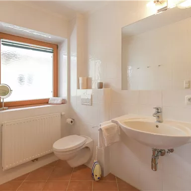 A modern bathroom with white tiles and a large window. There is a sink, a toilet, and a heater.