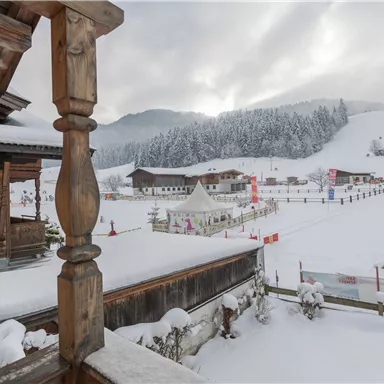A snowy winter landscape with a mountain backdrop and traditional wooden houses. In the foreground, a ski area is visible with a tent and many snow-covered trees.