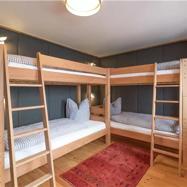 A cozy room with a wooden bunk bed and two mattresses. The floor is made of wood, and a red carpet adds extra coziness.