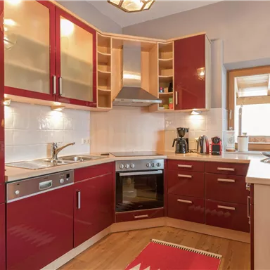 A modern kitchen with red cabinets and a wooden floor. It is well-equipped with appliances and has large windows for plenty of natural light.