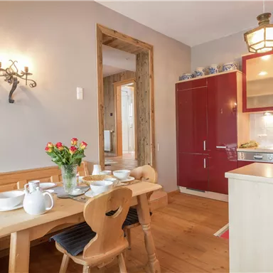 A cozy kitchen with a wooden table and chairs. The walls are painted in soft colors, and there is a red refrigerator as well as fresh flowers on the table.