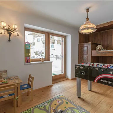 A cozy room with wooden flooring and a window that lets in daylight. On the left side, there is a small table, and on the right side, there is a table football table.