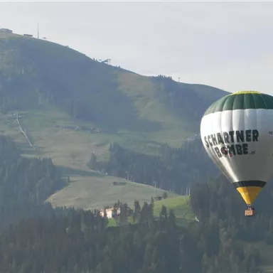 Ein Heißluftballon schwebt über einer grünen Berglandschaft. Im Hintergrund sind sanfte Hügel und Bäume zu sehen.