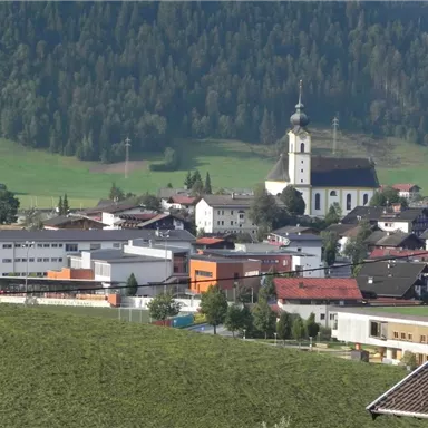 Eine malerische Aussicht auf ein kleines Dorf mit einer Kirche und umliegenden Bergen. Die Gebäude sind umgeben von grünen Wiesen und Bäumen.