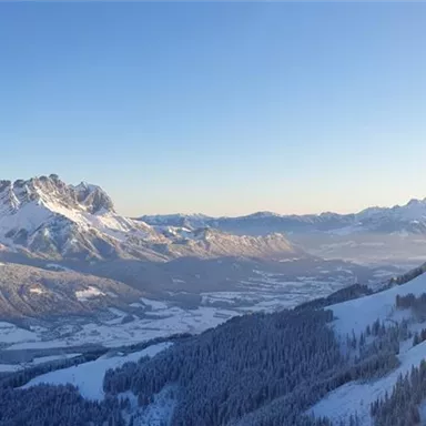 Eine atemberaubende Winterlandschaft mit schneebedeckten Bergen und einem klaren blauen Himmel. Im Vordergrund ist eine Seilbahn sichtbar, die ins Gebirge führt.