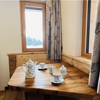 Ein gemütlicher Tisch aus Holz mit einer Teekanne und Tassen. Durch das Fenster sieht man eine verschneite Landschaft.