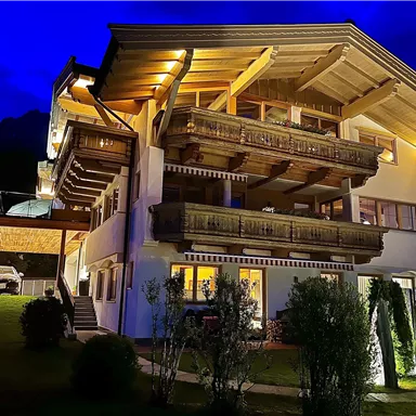A beautiful chalet with wooden cladding and balconies, illuminated at night. The garden is well-maintained and offers an inviting atmosphere.