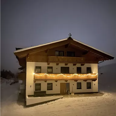 A cozy house in the snow, illuminated by warm lights. The sky is dark and there is a calm winter atmosphere in the air.
