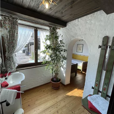 A cozy room with wooden floors and a large window. There are plants and ski equipment hanging on the wall.