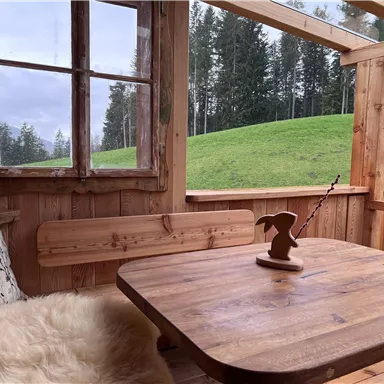 Ein gemütlicher Holzraum mit einem Tisch und einer Bank. Durch das Fenster blickt man auf eine grüne Wiese und Bäume.