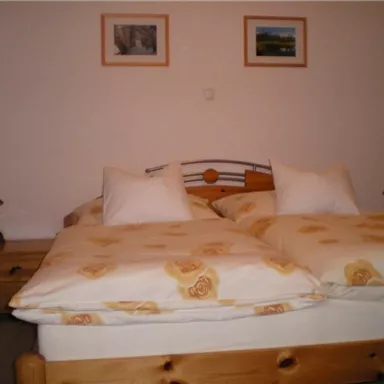 Ein gemütliches Schlafzimmer mit einem Doppelbett und Kissen. An den Wänden hängen zwei Bilder, und es gibt einen Nachttisch mit einer Lampe.