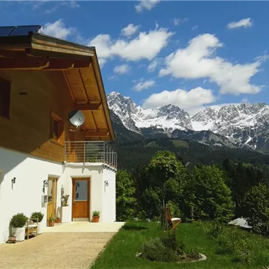 A modern house with a wooden facade and large windows. In the background, impressive snow-covered mountains and a blue sky can be seen.