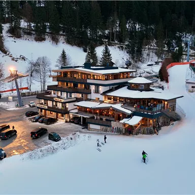 A modern mountain hotel in a snowy landscape with many fir trees. Skiers enjoy the slope in front of the hotel and several cars are parked in the parking lot.