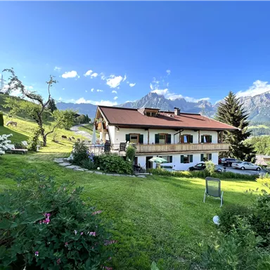 Ein schönes Haus in der Natur mit einem gepflegten Garten. Im Hintergrund sind Berge und ein klarer Himmel zu sehen.