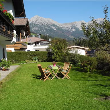 Eine schöne Grünfläche mit zwei Liegestühlen und einem Tisch, umgeben von Bäumen und Blumen. Im Hintergrund sind beeindruckende Berge zu sehen.