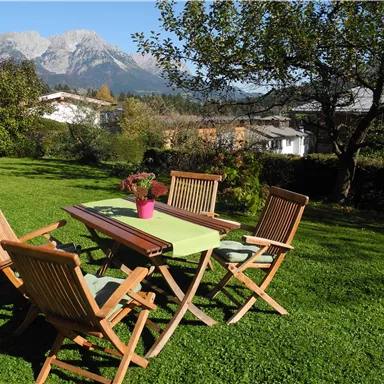 Ein schöner Garten mit einem Tisch und vier Stühlen. Die Berge sind im Hintergrund sichtbar und die Sonne scheint.