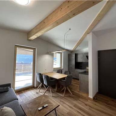 Ein modernes Wohnzimmer mit Holzbalken und großen Fenstern. Der Raum ist hell und gemütlich, mit einem Esstisch und einer komfortablen Sitzecke.