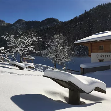 Eine verschneite Winterlandschaft mit einem gemütlichen Holzhaus. Im Vordergrund sind zwei Bänke und ein Spielplatz zu sehen, umgeben von schneebedeckten Bäumen.