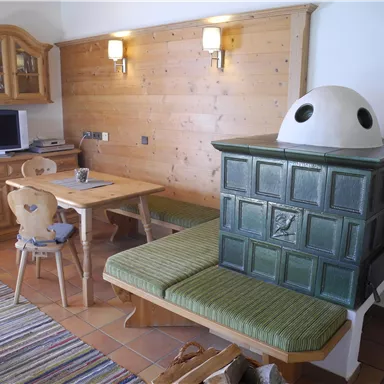 Ein gemütlicher Wohnraum mit Holzverkleidung und einem traditionellen Kachelofen. Ein Essbereich mit einem Tisch und Stühlen ergänzt die warme Atmosphäre.