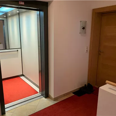 A modern elevator in a bright hallway with a red carpet. On the right side, a wooden and glass door is visible.