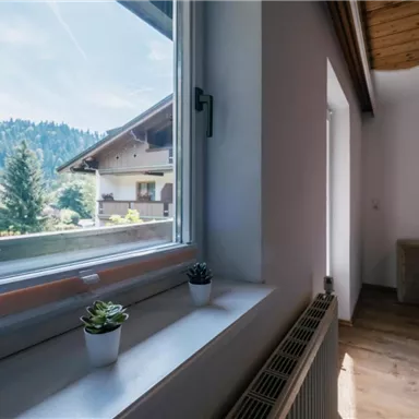 Ein helles Zimmer mit Blick auf die Berge. Auf dem Fensterbrett stehen zwei kleine Pflanzen und die Wände sind in sanften Farben gehalten.