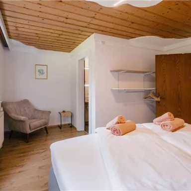 Ein helles Schlafzimmer mit einem Doppelbett und zwei Handtüchern. Im Hintergrund steht ein bequemer Sessel und ein kleiner Tisch.