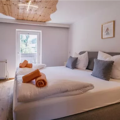 Ein modernes Schlafzimmer mit einem großen Bett, dekorierten Kissen und frisch gefalteten Handtüchern. Das Zimmer hat große Fenster und warmes Holzdesign.