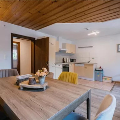 Eine gemütliche Wohnküche mit einem großen Tisch und modernen Stühlen. Der Raum hat helle Wände und Holzdecken, was eine warme Atmosphäre schafft.