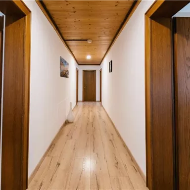 Ein heller Flur mit Holzpaneelen und zwei offenen Türen. An den Wänden hängen Bilder, und der Boden ist aus Holz.