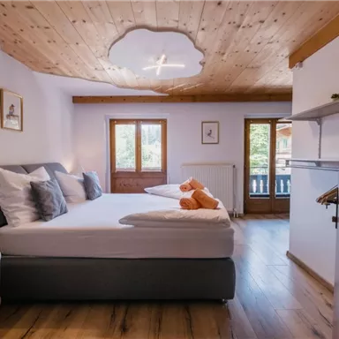 Ein gemütliches Schlafzimmer mit einem modernen Bett und einer warmen Holzdecke. Große Fenster sorgen für viel Tageslicht und Blick auf die Umgebung.